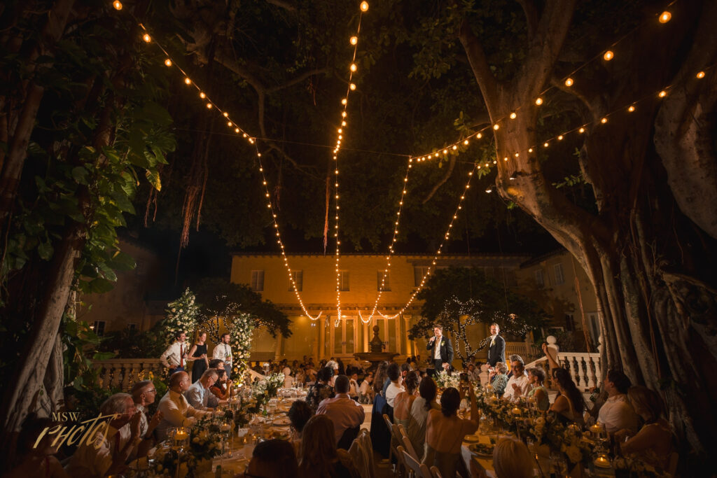 Wide angle scene of wedding toasts under banyan trees at night.